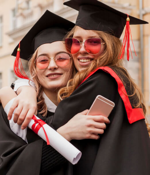 Two female graduates in caps and gowns celebrate their achievement. They are hugging, smiling, and holding diplomas.