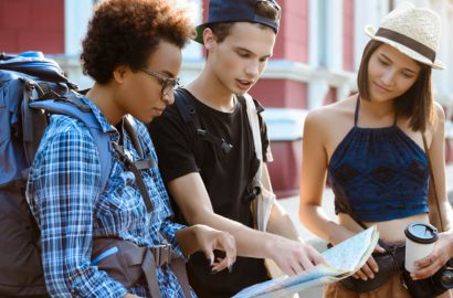 Three young travelers are examining a map. One has a backpack and plaid shirt, another wears a black cap, and the third has a hat and holds a coffee cup.