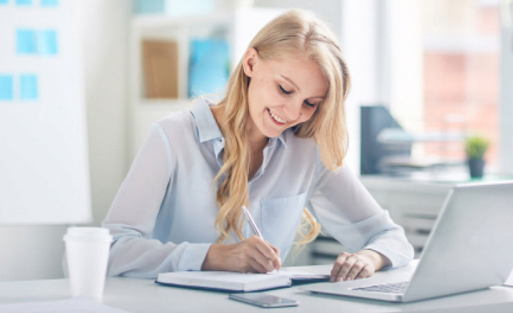 A professional woman, smiling and writing in a notebook, sits at her desk with a laptop, smartphone, and coffee cup.