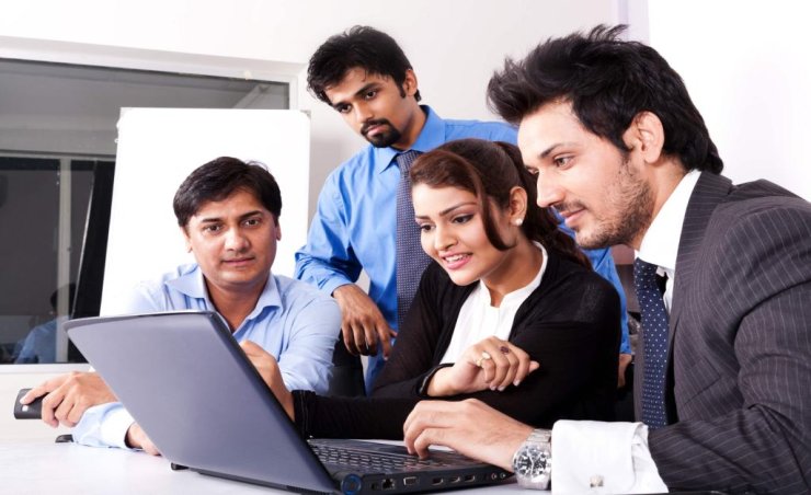 Four business professionals, two men and two women, are gathered around a laptop, engaged in a collaborative work session.