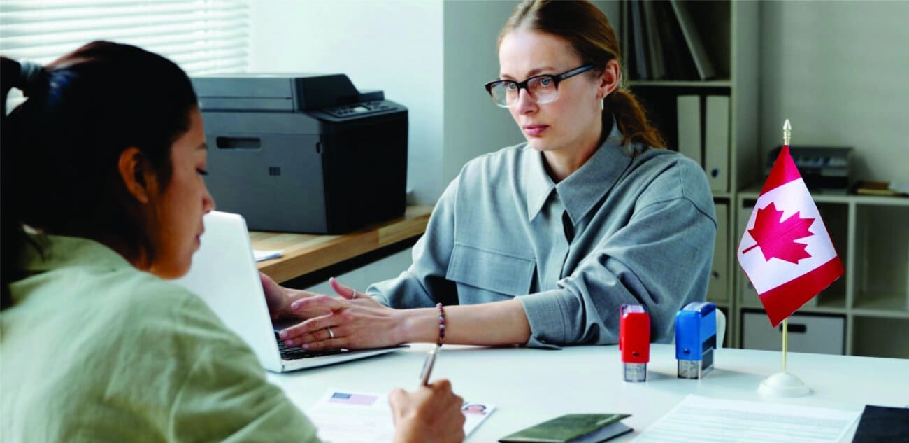 A woman in glasses works on a laptop across from another woman in an office with a Canadian flag on the desk. Likely a visa or immigration meeting.