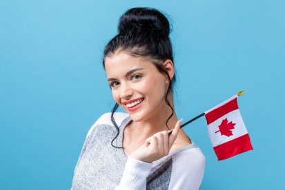 A smiling woman holds a small Canadian flag against a blue background. She is wearing a casual grey and white sweater, suggesting pride or celebration of Canada