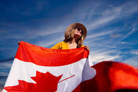 A woman is standing outdoors, holding a large Canadian flag. She is wearing a hat and sunglasses, looking upward, with a clear blue sky in the background.